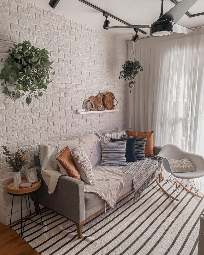 Sala boho com parede de tijolinhos, sofá cinza, almofadas coloridas e mantas