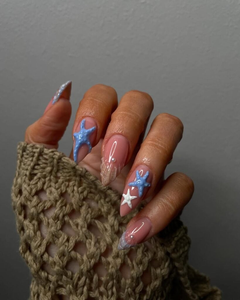 Mão de pele clara, com unha amendoada rosada com conchas e decorações que remetem ao mar