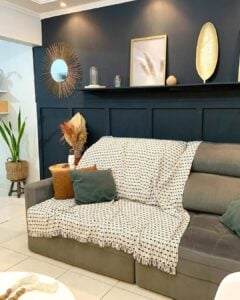 Sala com sofá cinza, manta clara e almofadas, parede escura com objetos decorativos