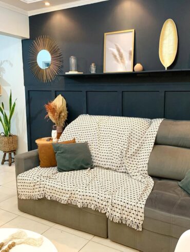 Sala com sofá cinza, manta clara e almofadas, parede escura com objetos decorativos