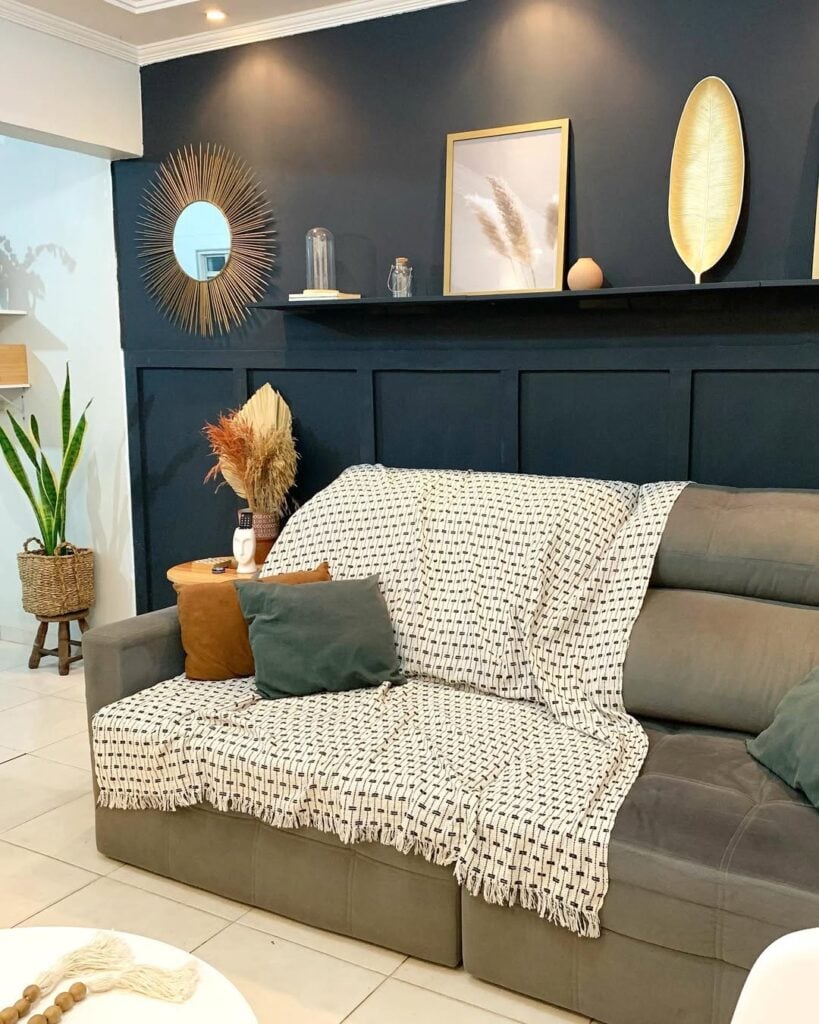 Sala com sofá cinza, manta clara e almofadas, parede escura com objetos decorativos