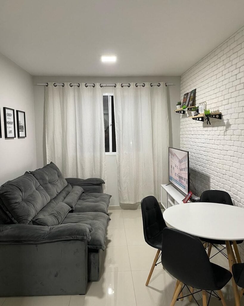 Sala com revestimento branco, sofá cinza em veludo e mesa redonda com cadeiras pretas