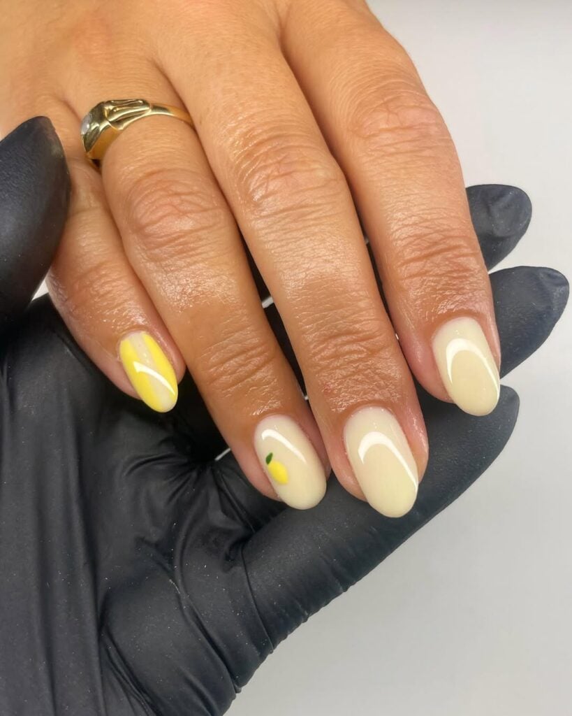 Mão de pele clara, com anel em dourado e unha amendoada amarelo suave com limão para decorar