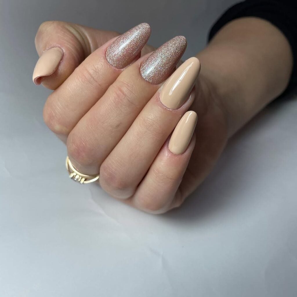 Mão de pele clara, com anel em dourado e unha amendoada bege com glitter champanhe