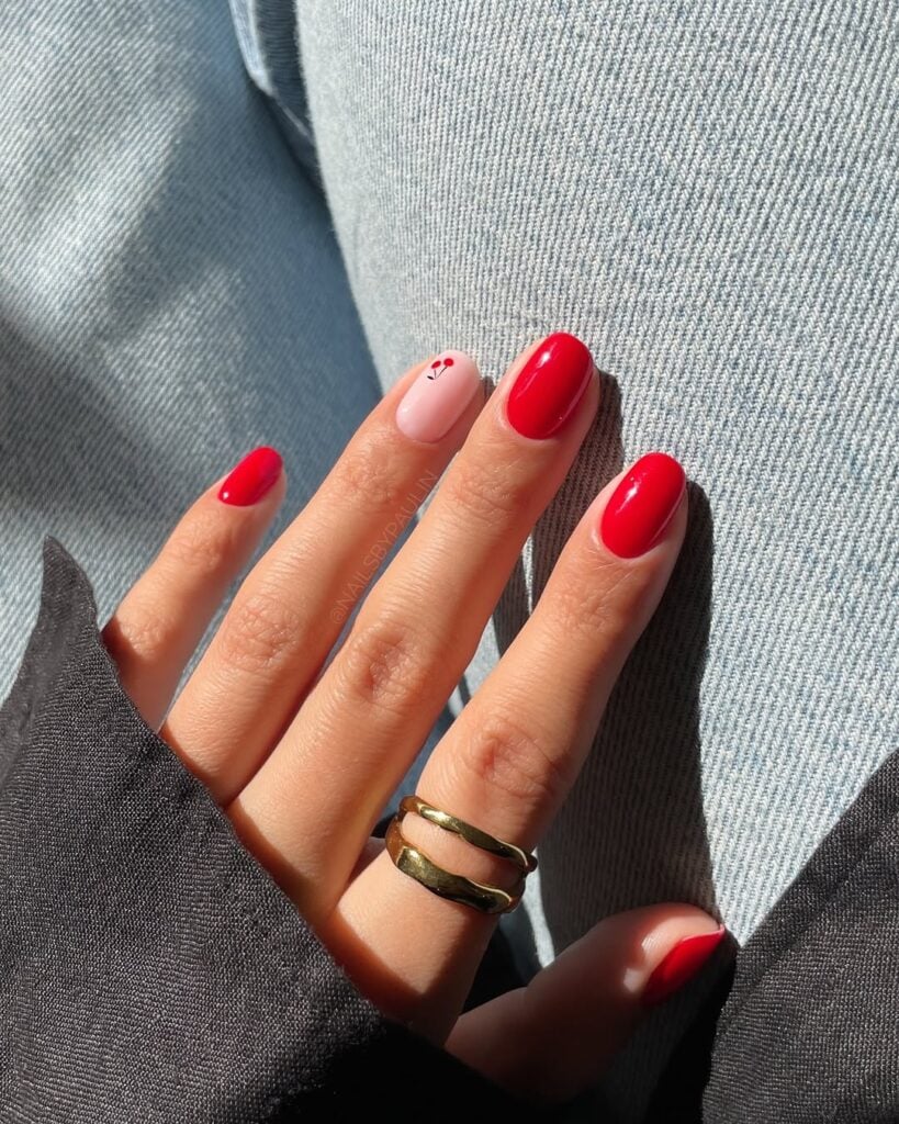 Mão de pele clara, com anel em dourado e unha squoval em vermelho e com cereja
