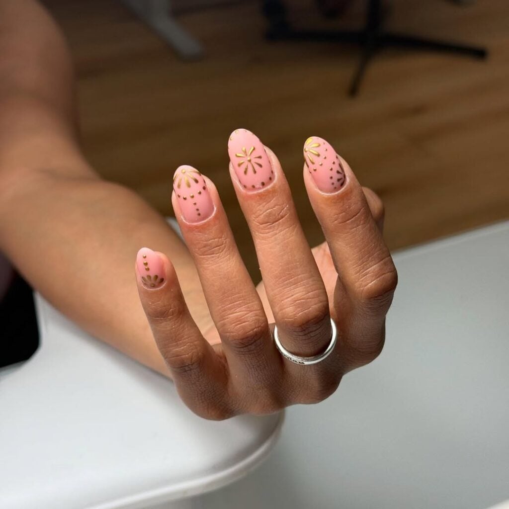 Mão de pele clara, com anel em prata e unha redonda rosada com decorações em dourado 