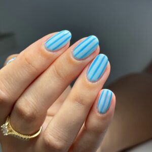 Mão de pele clara, com anel em dourado e unha redonda azul-claro com listas azul-escuro