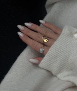Mão de pele clara, com anéis em dourado e unha amendoada perolada com strass