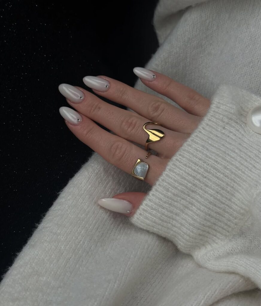 Mão de pele clara, com anéis em dourado e unha amendoada perolada com strass
