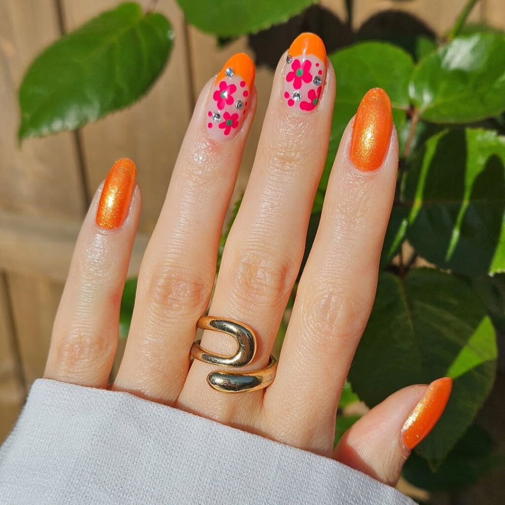 Mãos de pele clara, com anel em dourado e unha amendoada laranja brilhante, com inglesinha flores em rosa e strass