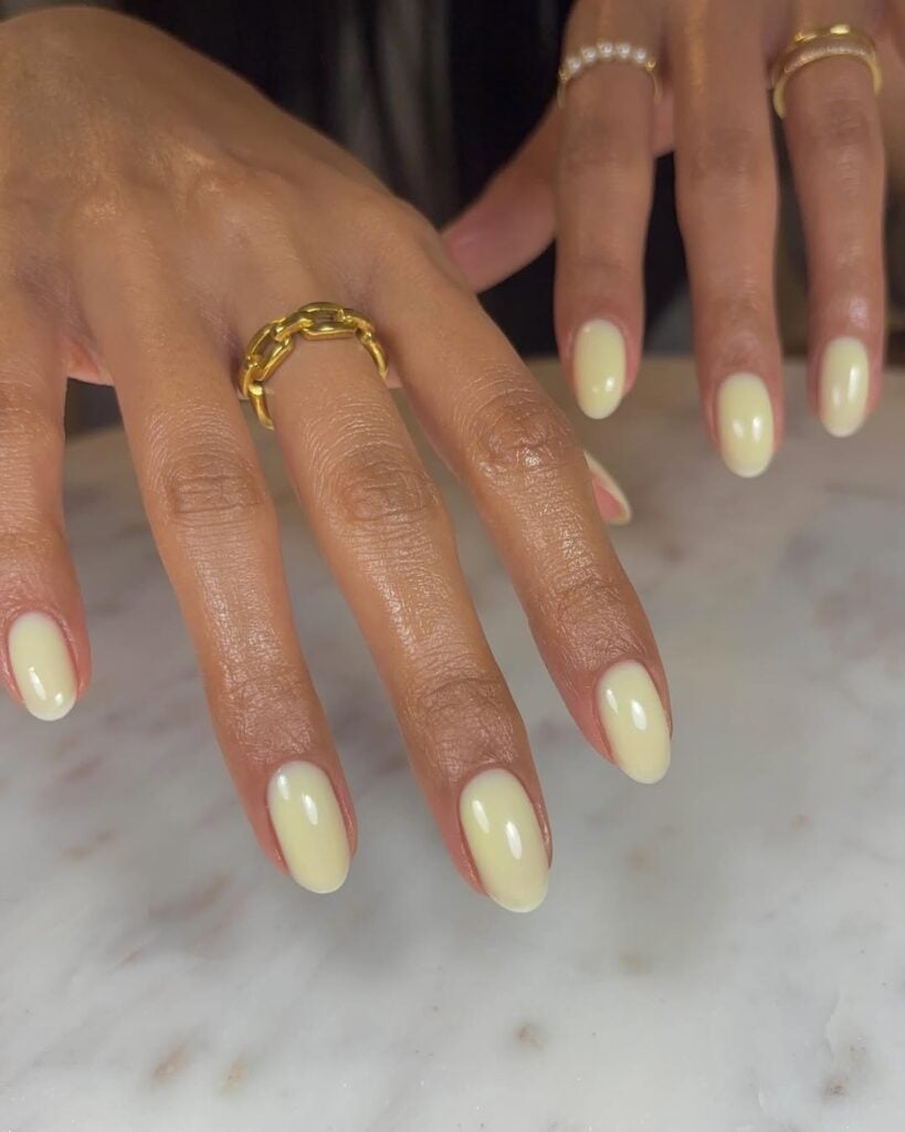 Mãos de pele clara, com anéis em dourado e unha amendoada amarelo claro