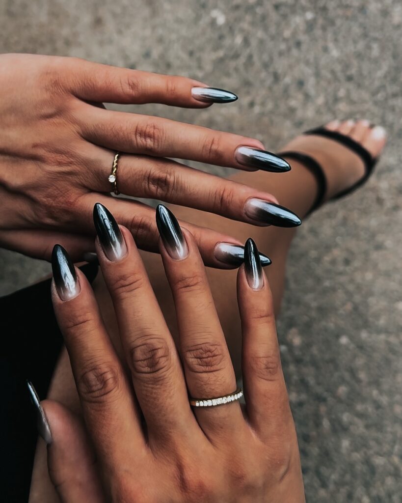Mãos de pele clara, com anéis em dourado e unha amendoada ombré, com esmalte preto e acabamento cromado
