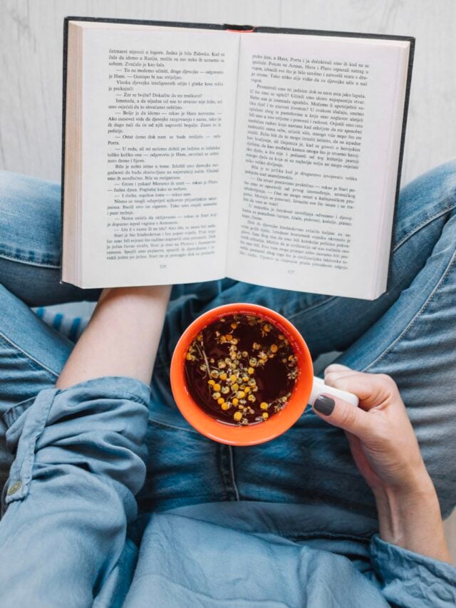 Mulher sentada de calça jeans e camisa azul com livro e uma caneca de chá nas mãos