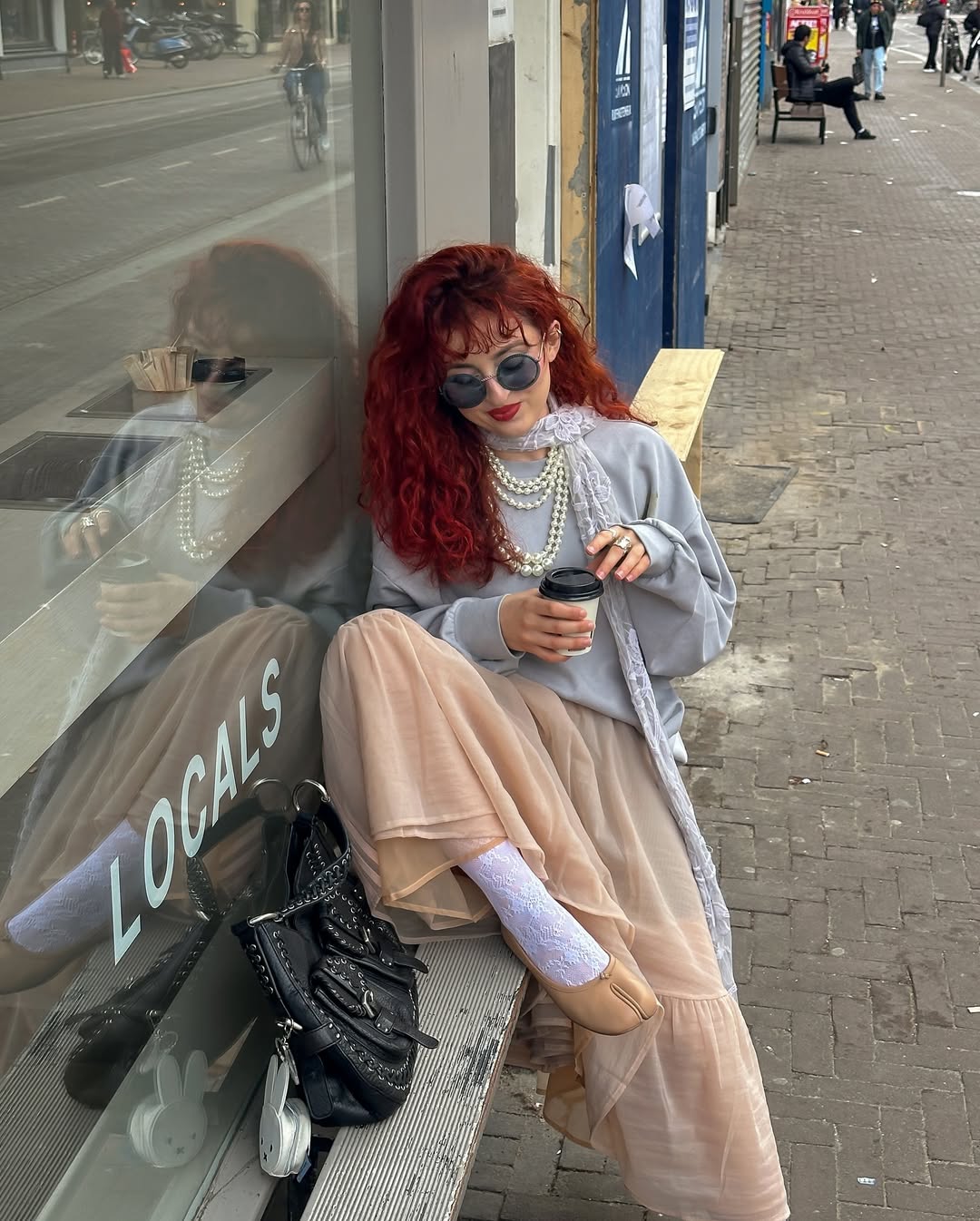 mulher jovem de cabelos vermelhos com look maximalista. Ela usa suéter, colar de pérolas, saia, meia e sapato pé de vaca.
