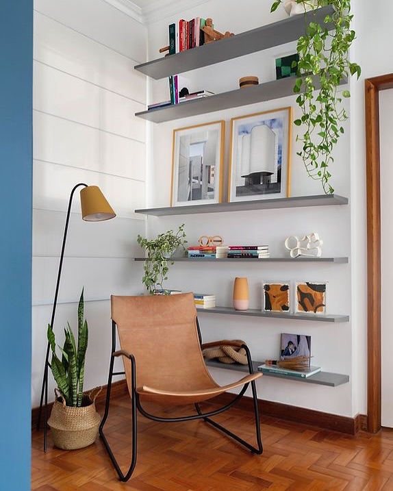 Ambiente com poltrona de metal preto com assento marrom, prateleiras cinza com objetos decorativos e livros