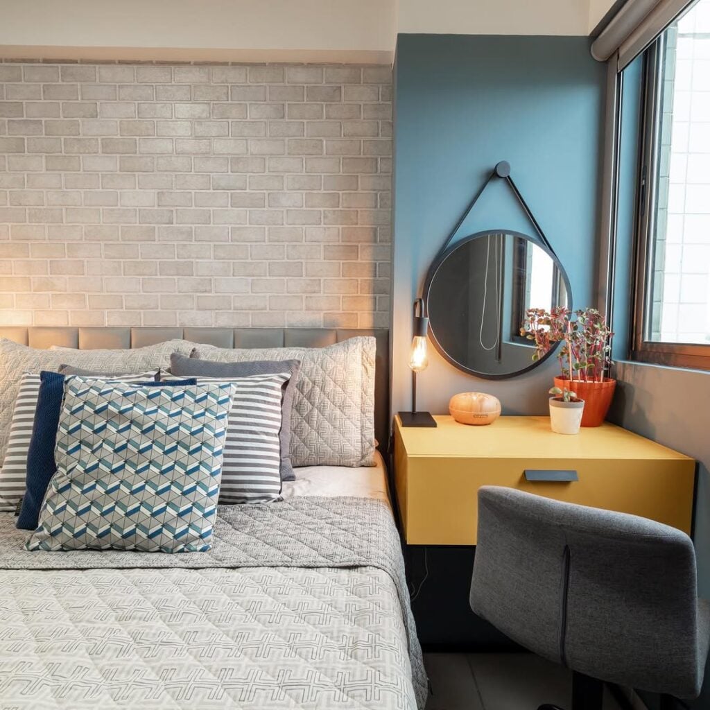 Quarto com papel de parede de tijolinhos cinza e cantinho da make com parede azul e penteadeira em tom de amarelo