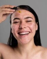 Mulher de pele clara e acneica, com cabelo escuro. Ela está aplicando algum sérum