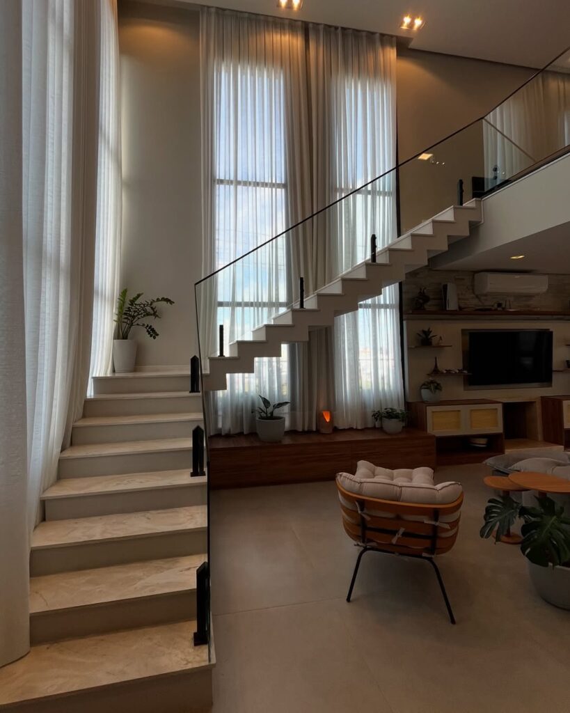 Sala de estar com pé-direito alto, escadaria e cortinas brancas