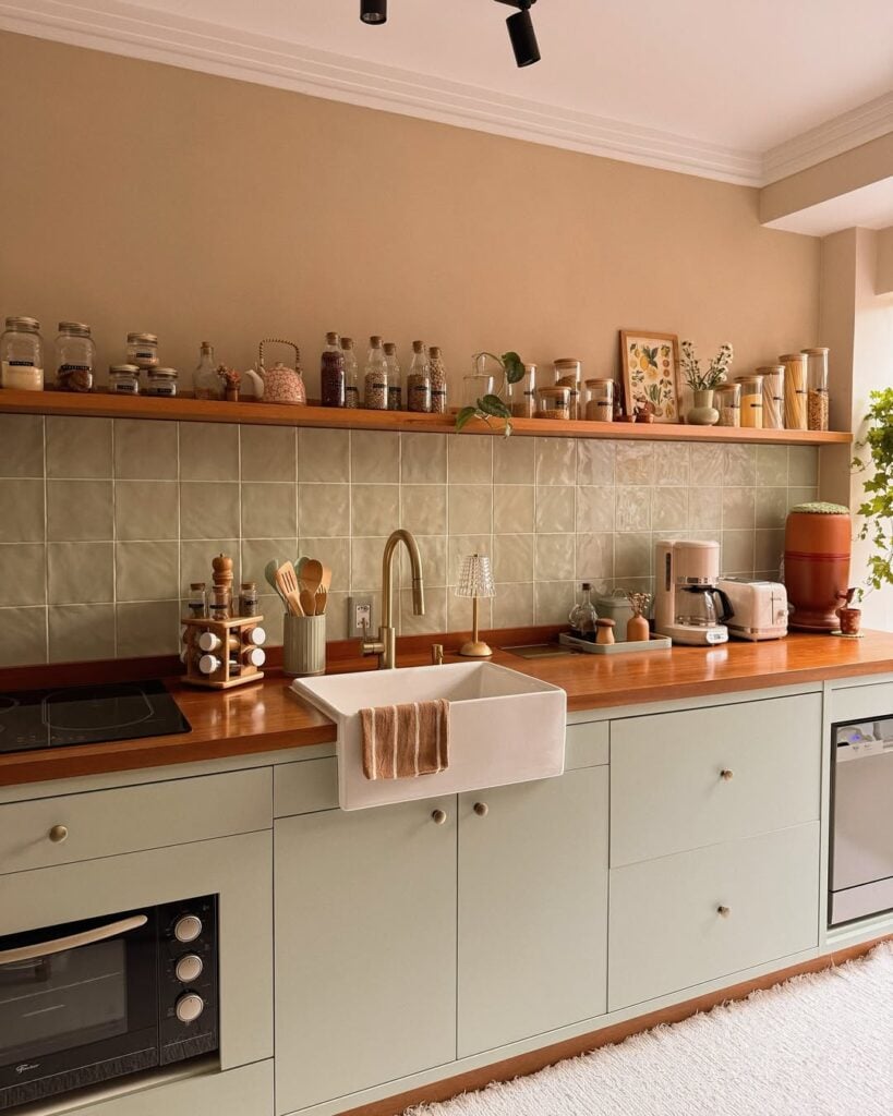 Cozinha com armário em tom de verde-menta com farm sink, bancada de madeira e prateleira com temperos