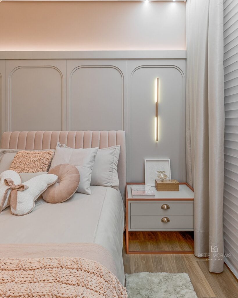 Quarto feminino com cabeceira aveludada rosa, mesa de cabeceira cinza-claro com puxadores em concha