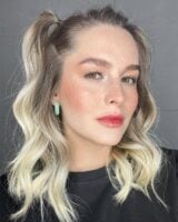 Mulher de pele clara, com blusa preta, cabelo loiro preso e lábios doll lips