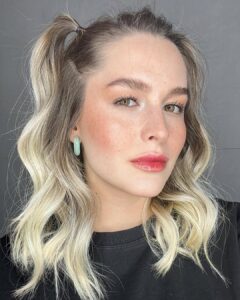 Mulher de pele clara, com blusa preta, cabelo loiro preso e lábios doll lips