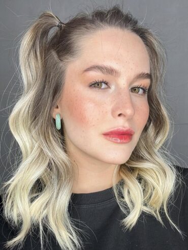 Mulher de pele clara, com blusa preta, cabelo loiro preso e lábios doll lips