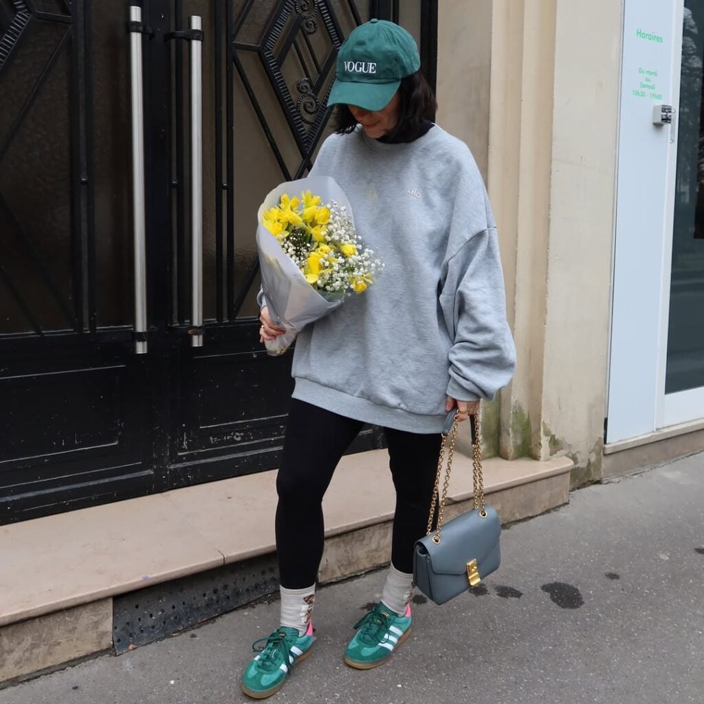 Mulher de pele clara, com blusão cinza, legging, tênis verde, boné verde e bolsa