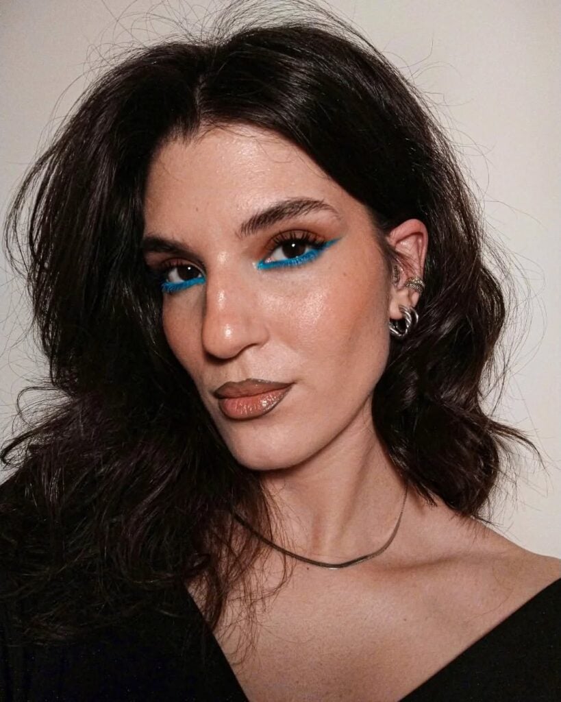 Mulher de pele clara, com blusa preta, acessórios em prata e maquiagem com lápis azul nos cílios inferiores