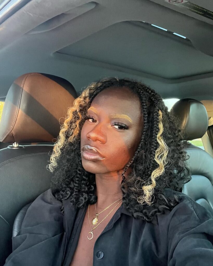 Mulher de pele preta, com cabelo cabelo crespo escuro com mechas loiras e com sobrancelha descolorida