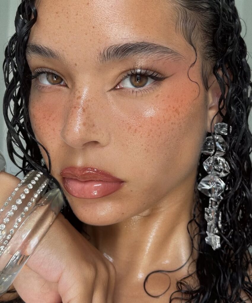 Mulher de pele preta clara, com cabelo cacheado escuro, acessórios transparentes e maquiagem clean