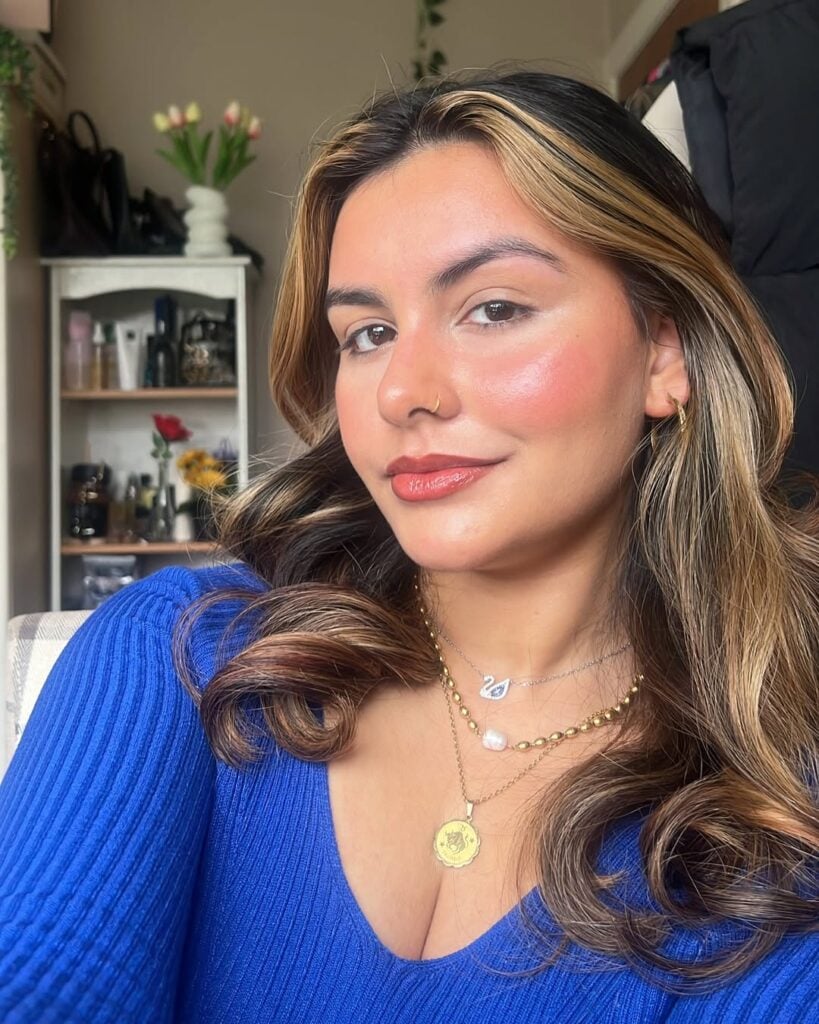 Mulher de pele clara, com cabelo iluminado, blusa azul, acessórios em dourado e maquiagem sem máscara de cílios