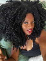 Mulher de pele preta, com blusa preta, cabelo crespo preto e maquiagem clean