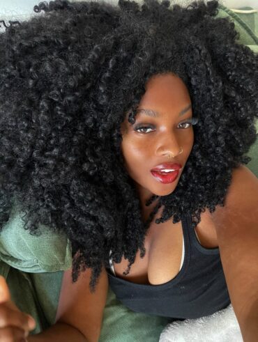 Mulher de pele preta, com blusa preta, cabelo crespo preto e maquiagem clean
