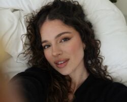 Mulher branca deitada com cabelo cacheado longo, usando maquiagem leve com gloss