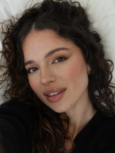 Mulher branca deitada com cabelo cacheado longo, usando maquiagem leve com gloss