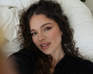 Mulher branca deitada com cabelo cacheado longo, usando maquiagem leve com gloss