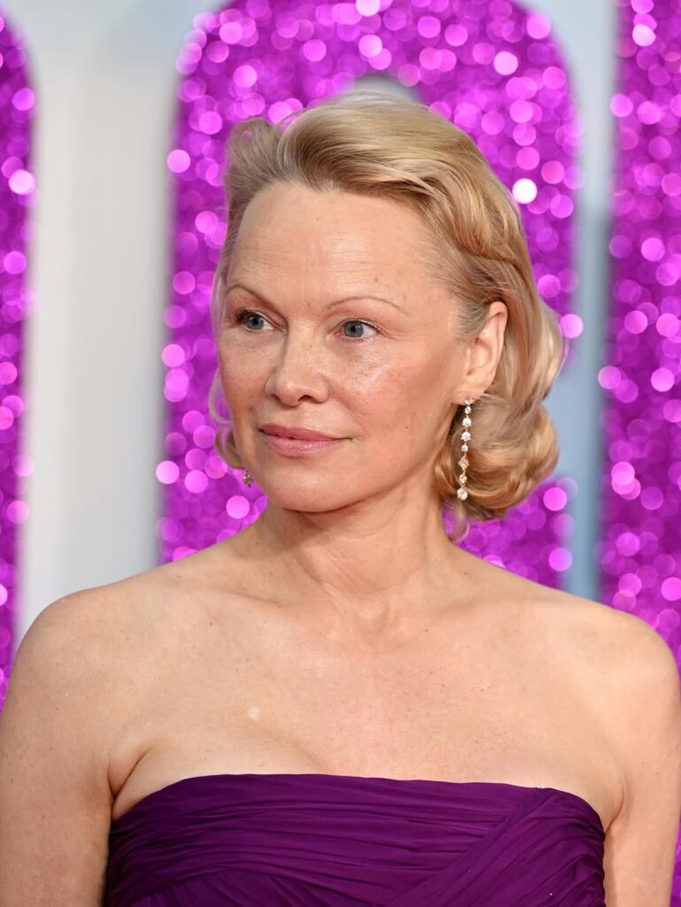 Pamela Anderson, com cabelo loiro, vestido roxo e sobrancelha fina
