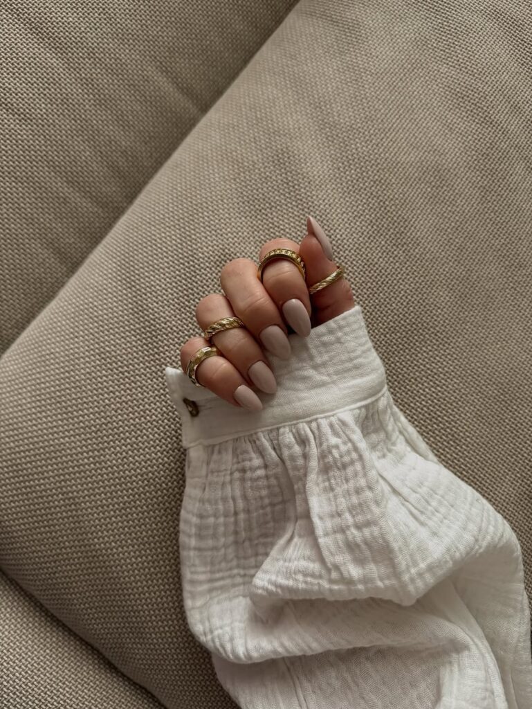 Mão de pele clara, com anéis em dourado e unha amendoada bege
