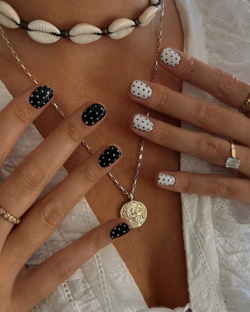 Mãos de pele clara, com acessórios em dourado e unha curta. Unhas decoradas com poá em preto e branco.