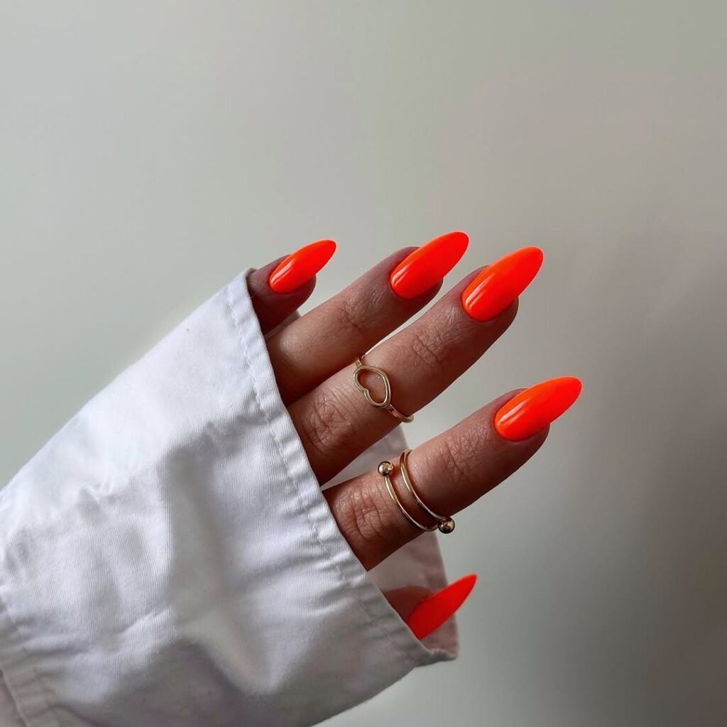 Mão de pele clara, com anéis em dourado e unha amendoada laranja