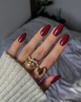 Mão de pele clara, com anéis em dourado e unha amendoada vinho
