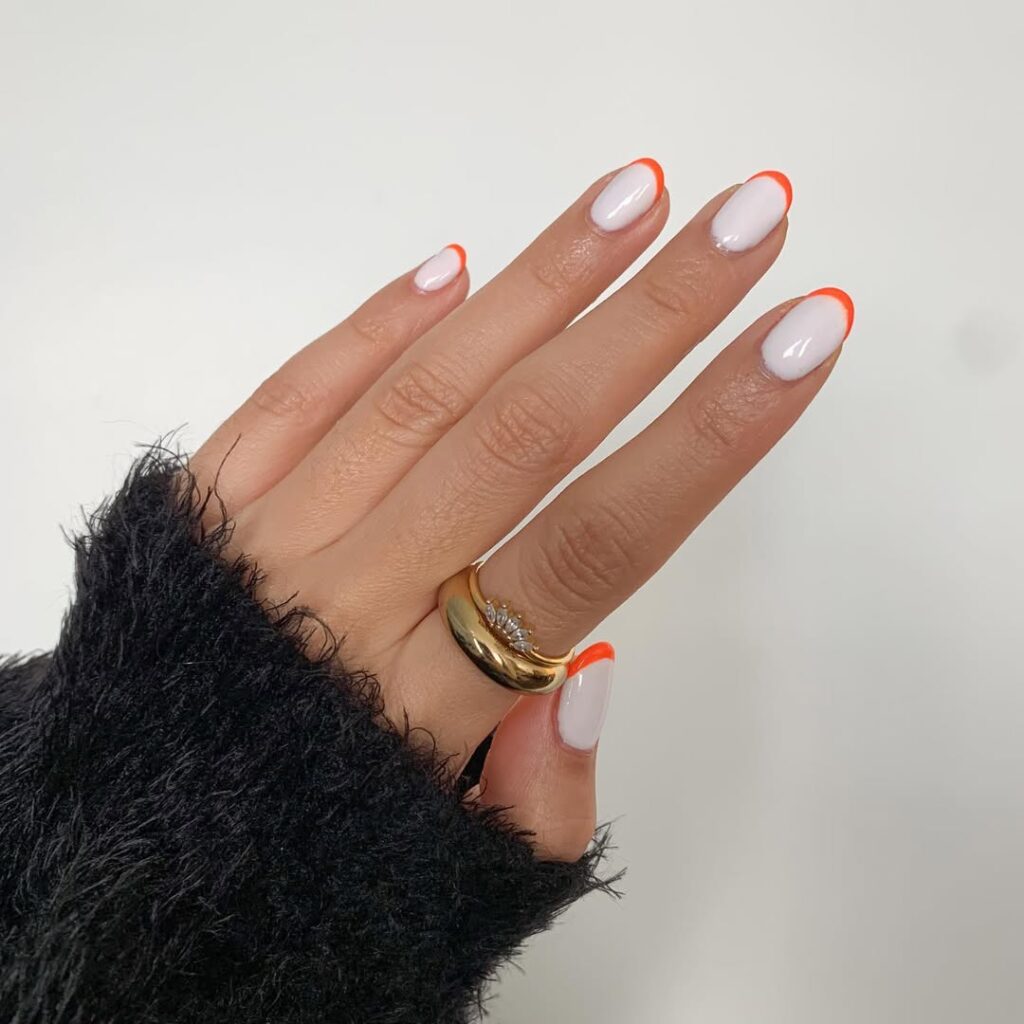 Mão de pele clara, com anel em dourado e unha amendoada branca com francesinha laranja