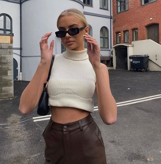 Mulher jovem e magra com pele clara. Ela usa blusa clara, calça marrom e óculos vintage preto