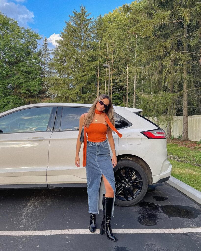 Mulher jovem na frente de um carro posando para foto. Ela usa blusa laranja sem alças, saia longa jeans e bota de cano longo preta