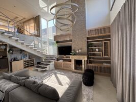 Sala de estar ampla com cores neutras, sofá cinza, escada e detalhes amadeirados