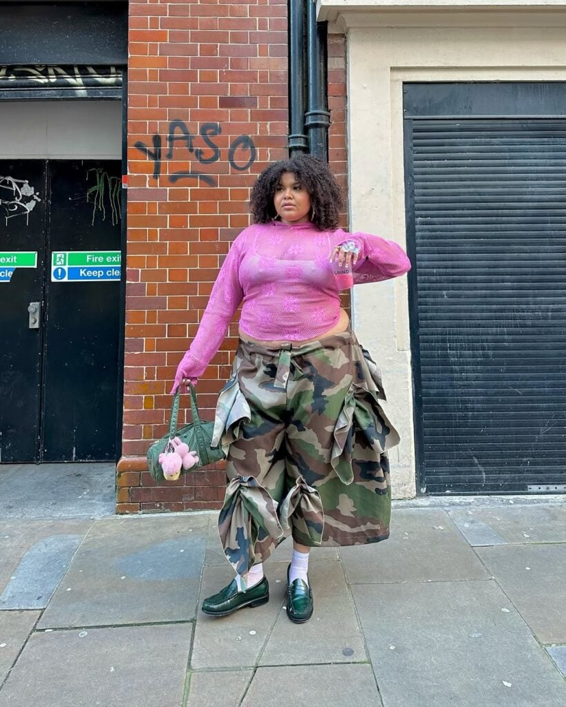 Mulher de pele preta clara, com calça camuflada, blusa de renda rosa, bolsa e mocassim verde