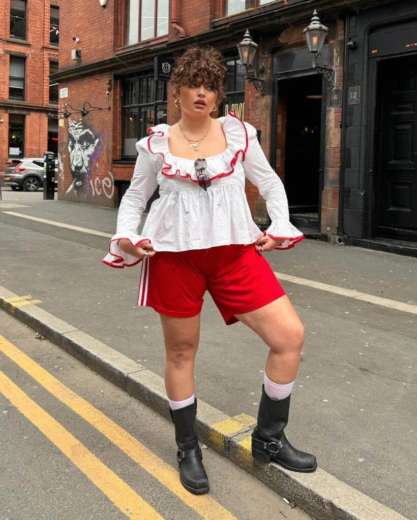 mulher jovem mid size com blusa bata manga longa branca, bermuda vermelha e bota biker preta