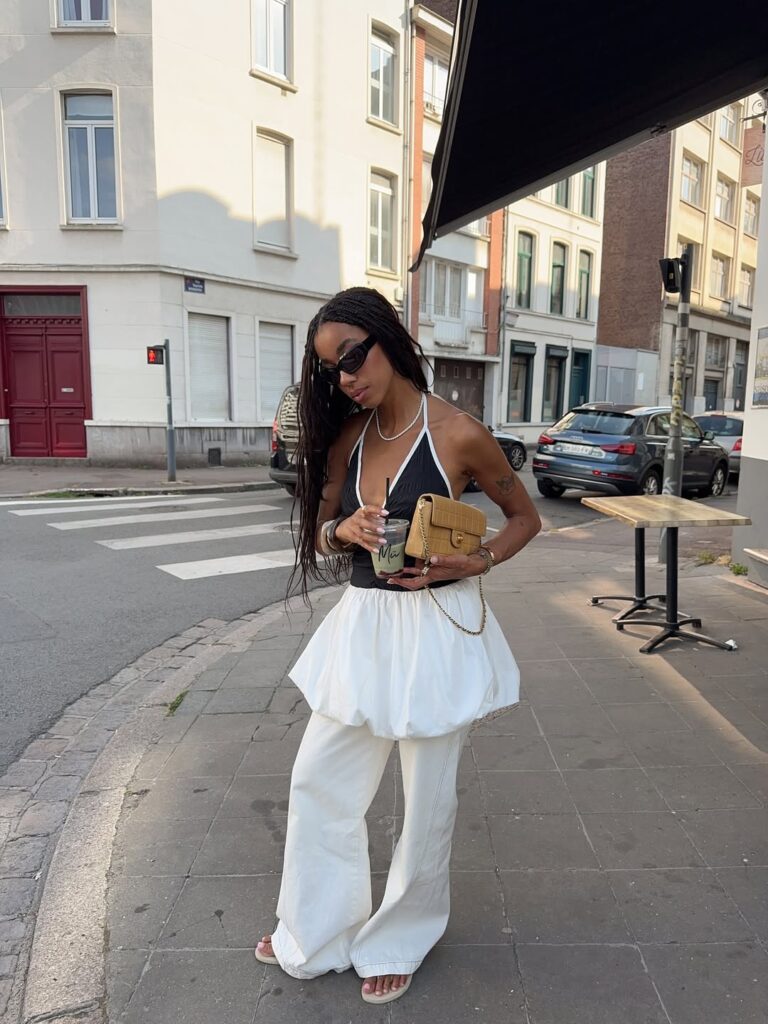 Mulher de pele preta, com cabelo trançado, regata preta, calça branca e chinelo de dedo