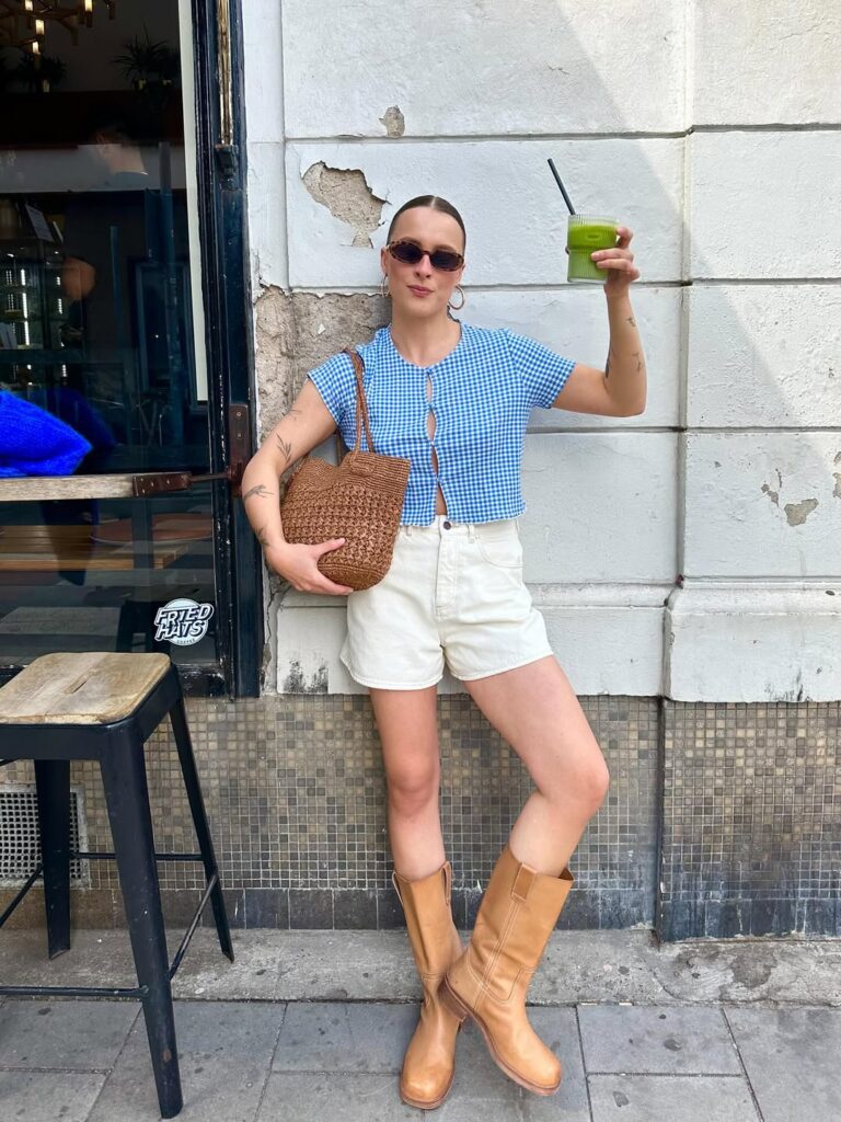 Mulher jovem com pele clara. Ela usa camiseta azul-claro, bermuda jeans e botas de cano médio